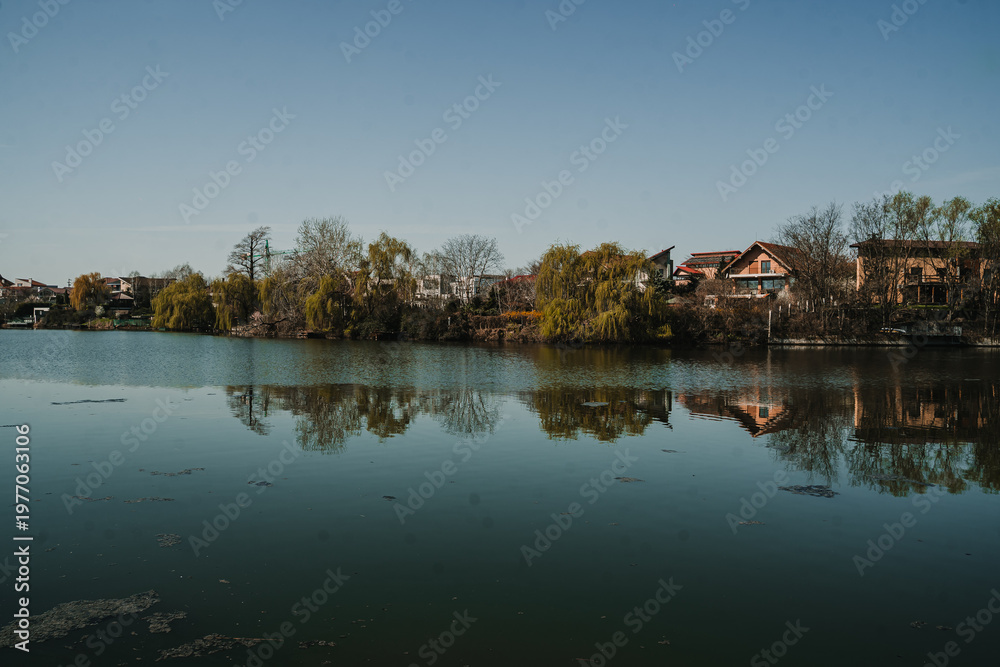 Fototapeta premium Peaceful lake landscape with reflections of houses and autumn trees in calm water. Quiet suburban scenery with natural reflections and soft light.