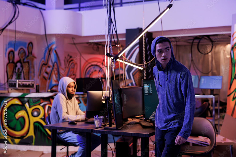 Fototapeta premium Suspicious hacker looking away after breaking into server and doing illegal activity. Young asian criminals in hood leaving abandoned warehouse after breaching database and stealing data