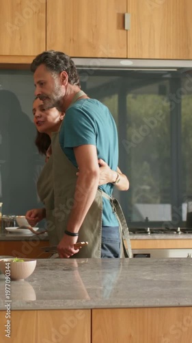 Vertical video: Hug releasing, couple moving to stove at kitchen island in aprons, stirring pan