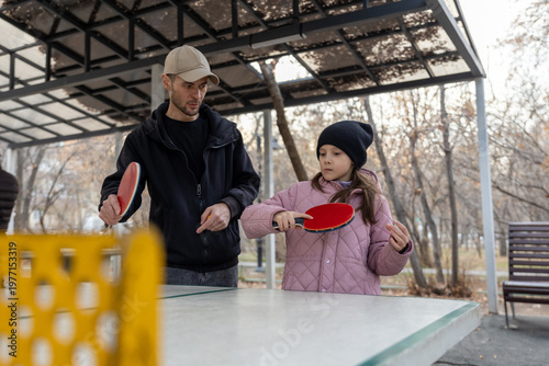 A father shows his daughter how to play ping pong at an outdoor sports ground under a canopy in a city park.