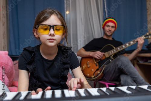 A young girl plays the piano in the foreground while her father plays an electric guitar in the background.