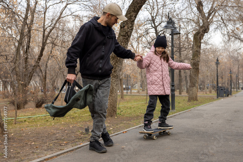 Man holding his daughter's hand to help her balance on a skateboard during a walk in the park.