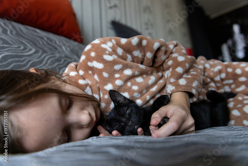 Close up of a young girl lying down and sleeping peacefully while holding a tiny black kitten in her arms.