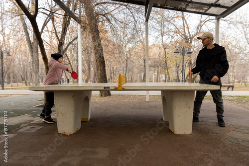 Full side view of a man and a girl playing ping pong on a concrete table surrounded by trees in a quiet outdoor setting.