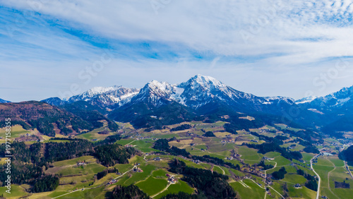 Aerial view of snow-capped mountains rise majestically over the lush green valley, creating a stunning contrast against the bright blue sky, Windischgarsten, Upper Austria, Austria.