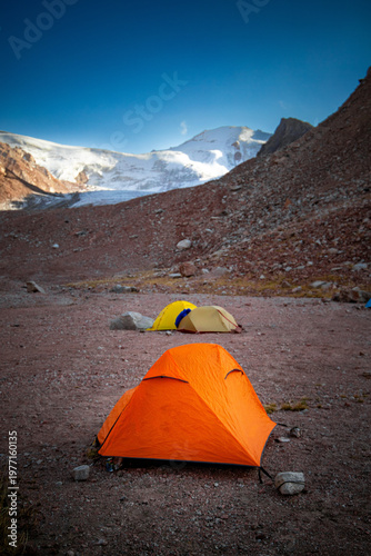 Orange and yellow tents pitched on a rocky mountain plateau with snow-capped peaks and glaciers in the background. Clear blue sky.