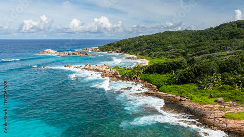 The turquoise ocean blending into the lush, green coastline with rocky formations. La Digue, Seychelles.