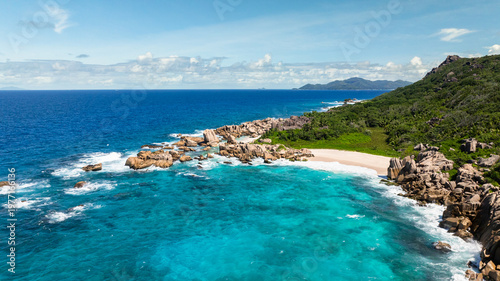Granite coastline with clear turquoise waters, white sandy beach, and vibrant greenery. Anse Marron beach. La Digue, Seychelles.