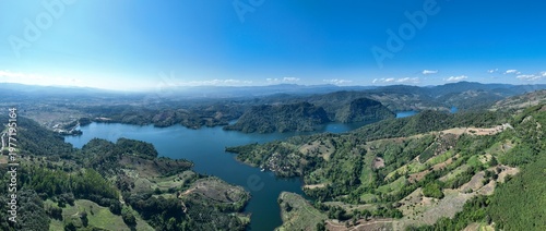 Vast Panoramic Landscape View Of A Winding Mountain Reservoir Surrounded By Lush Green Hills And A Small Rural Village Under A Clear Blue Sky