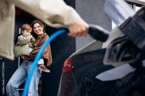 Family standing as father charges electric car at home, mother holding child. Embracing clean energy and ecological lifestyle