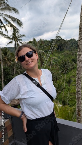 Woman smiles, waving joyfully from elevated wooden platform. Tropical green hills roll behind her, palm trees sway. She wears casual white shirt and black high-slit skirt