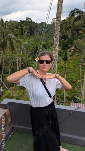 Woman smiles, waving joyfully from elevated wooden platform. Tropical green hills roll behind her, palm trees sway. She wears casual white shirt and black high-slit skirt