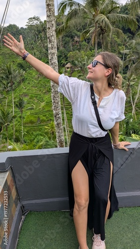 Woman smiles, waving joyfully from elevated wooden platform. Tropical green hills roll behind her, palm trees sway. She wears casual white shirt and black high-slit skirt