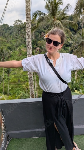 Woman smiles, waving joyfully from elevated wooden platform. Tropical green hills roll behind her, palm trees sway. She wears casual white shirt and black high-slit skirt