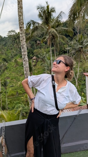 Woman smiles, waving joyfully from elevated wooden platform. Tropical green hills roll behind her, palm trees sway. She wears casual white shirt and black high-slit skirt