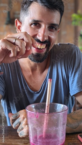 Young man drinking fresh tropical smoothie at bar, healthy lifestyle and wellness concept, smiling bearded athlete enjoying juice with straw, natural fruit breakfast, positive energy and confidence