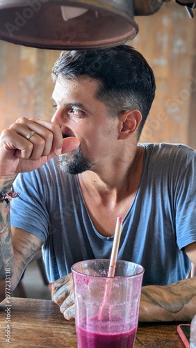 Young man drinking fresh tropical smoothie at bar, healthy lifestyle and wellness concept, smiling bearded athlete enjoying juice with straw, natural fruit breakfast, positive energy and confidence