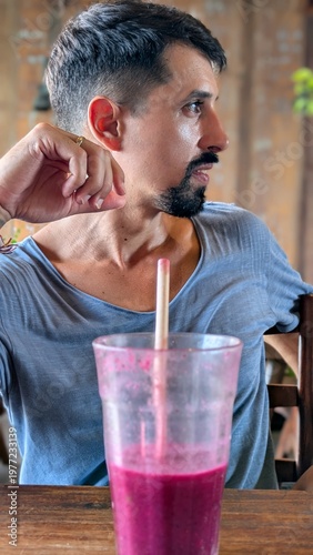 Young man drinking fresh tropical smoothie at bar, healthy lifestyle and wellness concept, smiling bearded athlete enjoying juice with straw, natural fruit breakfast, positive energy and confidence