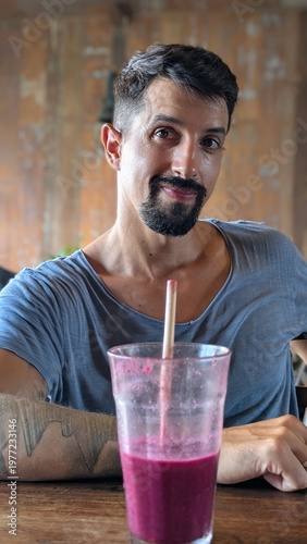Young man drinking fresh tropical smoothie at bar, healthy lifestyle and wellness concept, smiling bearded athlete enjoying juice with straw, natural fruit breakfast, positive energy and confidence