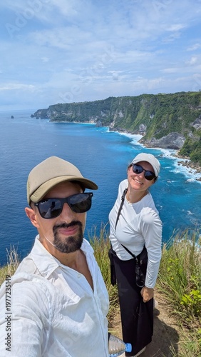Couple taking selfie at Manta Point cliff in Bali Indonesia, tropical paradise with turquoise ocean and panoramic seascape, friends enjoying iconic viewpoint, risky edge adventure with stunning waves 