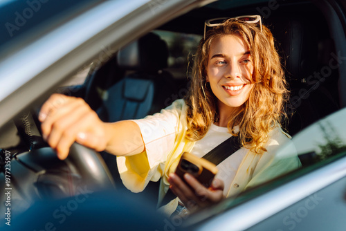 фотографія Young woman sits behind wheel of car, holding phone, on sunny day