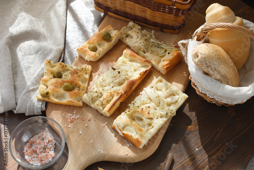 Assorted Italian focaccia slices with olives and onions on wooden board. High-angle shot features variety of artisan flatbreads with fresh bread rolls in wicker basket and pink salt. Professional food