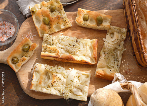 Traditional Italian focaccia assortment with green olives and onions on rustic wooden board. High-angle shot features artisan flatbread slices with pink Himalayan salt and fresh bread rolls in wicker