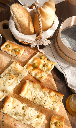 Artisan Italian focaccia slices with olives and onions on wooden board with traditional flour sieve. High-angle shot features variety of homemade breads pink Himalayan salt and wicker basket under