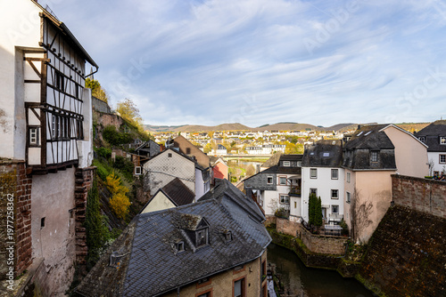 Scenic view of Saarburg with traditional houses along a narrow river, overlooking rooftops, historic architecture, and a peaceful valley landscape under a dramatic sky