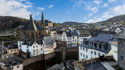 Panoramic view of Saarburg, Germany, featuring historic old town architecture with half-timbered houses, a riverside setting, and a traditional church, creating a picturesque European landscape under 