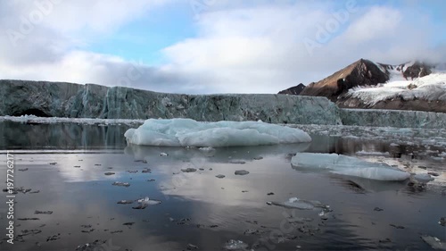 Glacial ice meets Arctic ocean in Svalbard. See icebergs floating serenely near massive glacier face. Clouds drift above snow-capped mountains in background, creating peaceful, frozen vista.