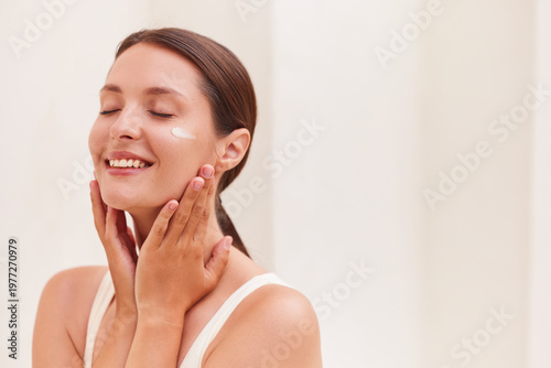 Natural beauty woman smiling joyfully with eyes closed and a cream swatch on her cheekbone, cradling her face with both hands during her skincare routine against a soft neutral background.