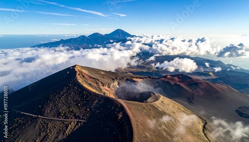 Elevated view of a volcanic crater with a winding path, partially obscured by clouds and other mountains in the distance