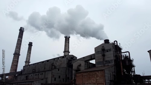 Industrial complex with tall smokestacks, buildings, and billowing smoke under an overcast sky