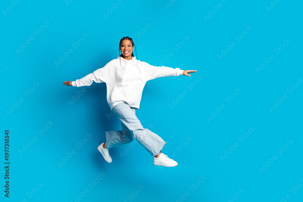 Fototapeta premium Young woman in white hoodie leaps with joy against blue background in a casual fashion photoshoot