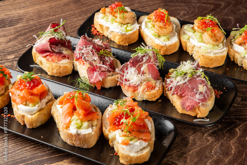 Bild auf Leinwand Assorted gourmet bruschettas with salmon, roast beef, and shrimp served on black plates over a dark wooden background