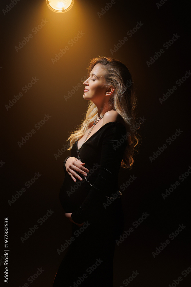 Fototapeta premium Pregnant woman in elegant black dress, artistic studio portrait with golden backlight