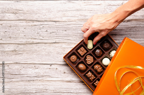 Female hand picking a chocolate bonbon from a box of assorted chocolate bonbons on wooden background.