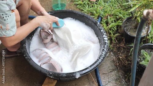 Person washing shoes by hand in a basin with soapy water outdoors. Suitable for cleaning, hygiene, and daily life themes.