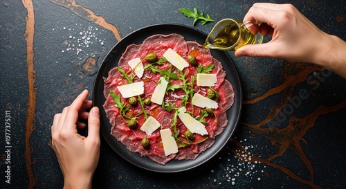 Beef carpaccio with parmesan and arugula being drizzled with olive oil on dark stone table