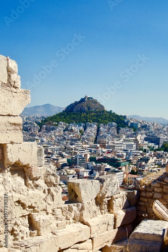 View of Athens, Greece with the prominent Lycabettus Hill in the background. The foreground features ancient stone ruins, part of the Acropolis. 