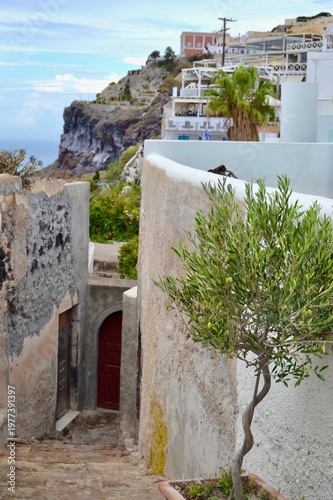 Narrow stone pathway to a cliff home in Santorini, Greece, flanked by traditional whitewashed buildings with rustic textures. 
