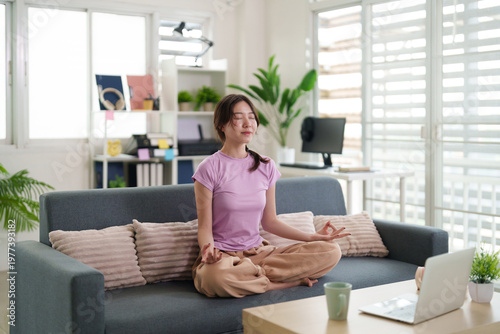 Asian adult woman practice meditation yoga for wellness mindfulness peace relax at modern home living room sofa stress relief healthy lifestyle balance