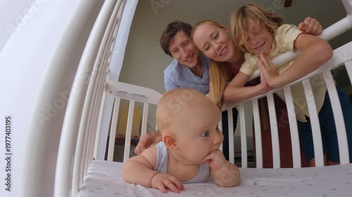 Family with mother, father and older brother gently looking at and touching a 3 month old baby lying in a crib. Tender bonding moment at home. Family love, newborn care and emotional connection