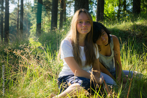 Two young girls sitting together in sunlit forest grass, one leaning on the other, natural lifestyle moment of friendship outdoors.