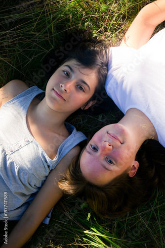 Two young girls lying on grass outdoors, looking at camera in warm sunlight, top view lifestyle moment of friendship.