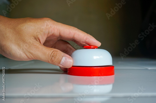 Close-up of a hand pressing a red and white service desk call bell on a table. Concept of customer service, assistance, urgency, order, or calling for help.