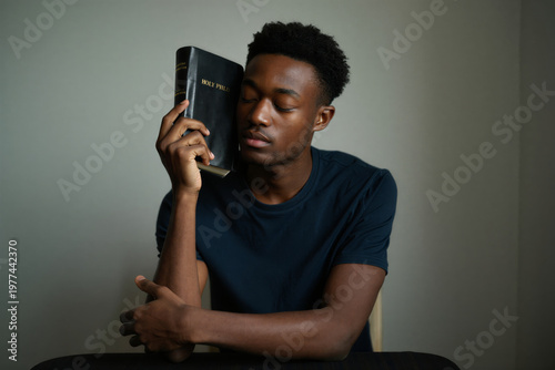 A young black man sitting and holding a Bible to his face with a contemplative expression