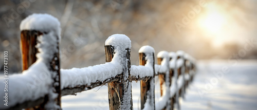 A rustic wooden fence covered in fresh snow glows under the warm morning sun. This peaceful winter landscape captures a serene, atmospheric mood with beautiful bokeh light.
