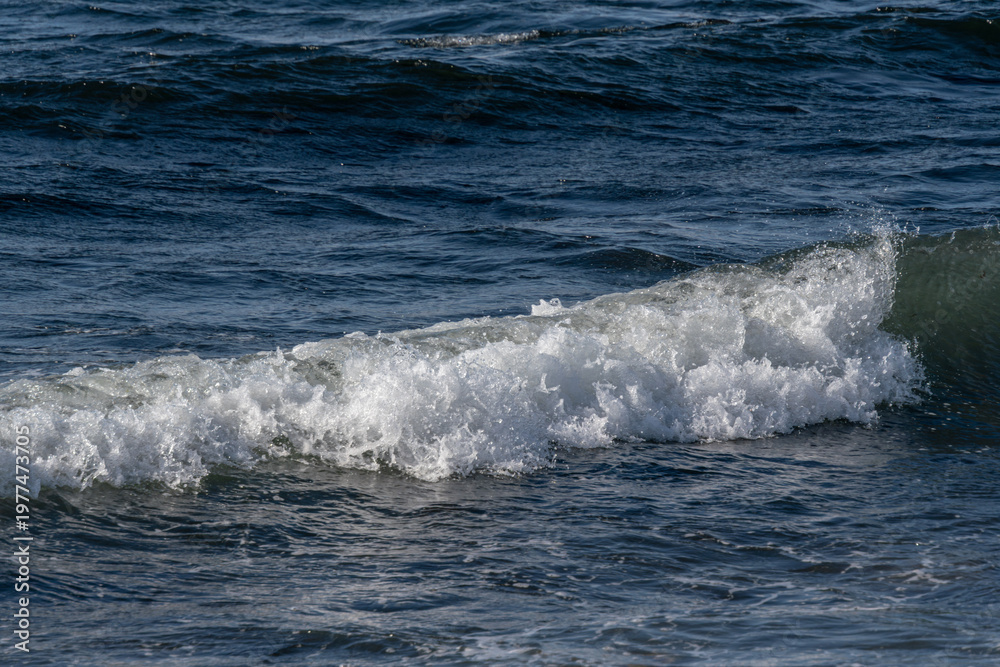 Obraz premium Ocean wave crashing on the shore with white foam and ripples, reflecting sunlight on the water surface, creating a serene coastal scene
